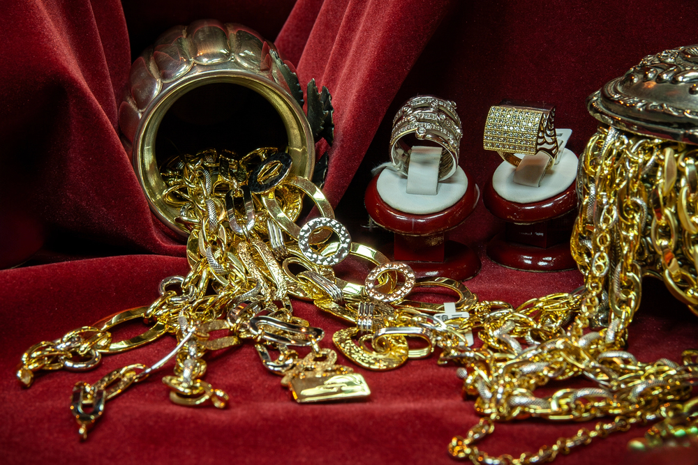 Gold necklaces, bracelets, and rings spill from ornate containers onto rich red velvet fabric, showcasing an array of sparkling jewellery pieces.