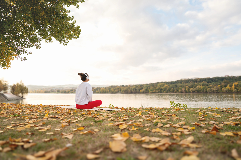 A person in a white jumper and red trousers sits cross-legged on grass covered with autumn leaves, facing a calm lake with trees in the background under a partly cloudy sky.