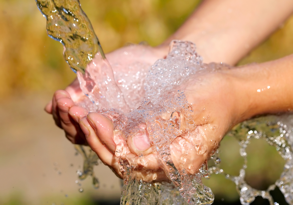 A close-up of two hands cupped together catching and holding flowing water outdoors, with a blurred natural background.