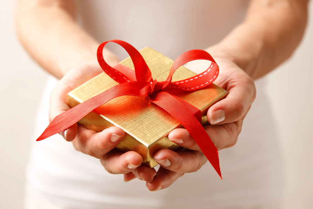 A person holding a small, gold-coloured gift box with a red ribbon and bow, offering it forward with both hands.