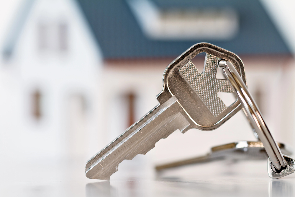 A close-up of a silver house key on a keyring, with a blurred image of a house in the background.