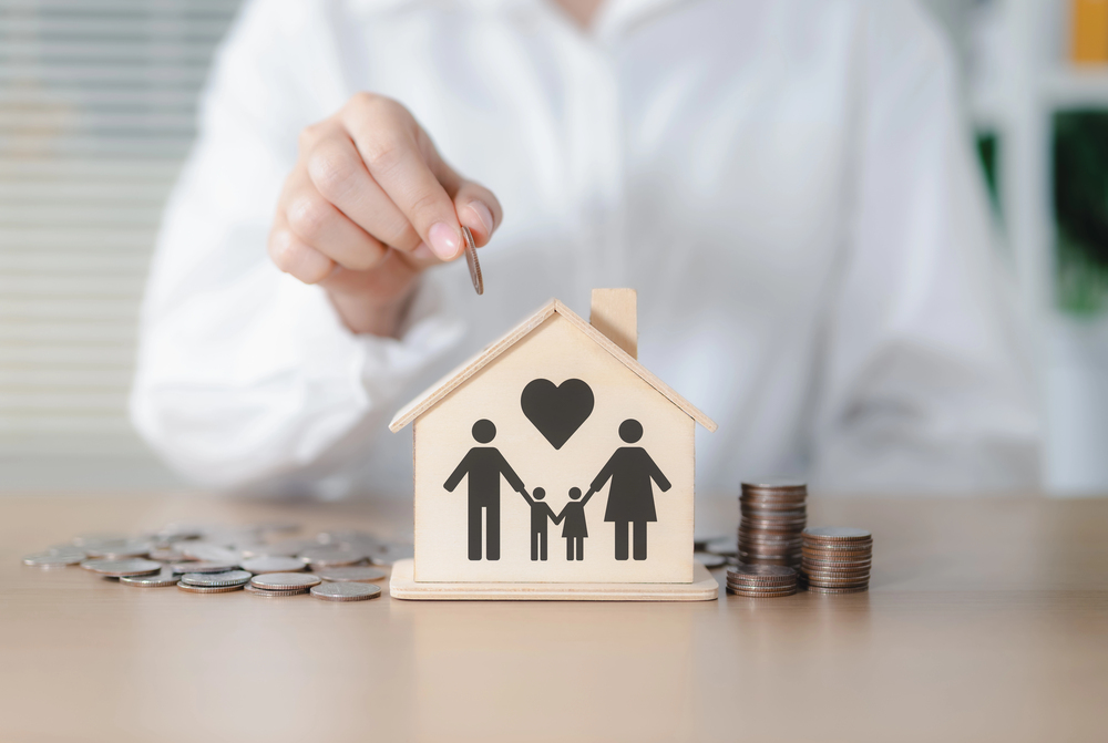 A person places a coin into a wooden house-shaped box with a family illustration and heart, symbolising saving money for family or home. Coins are stacked and scattered on the table.