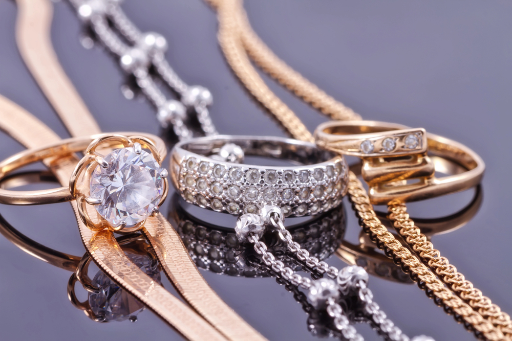 Assorted gold and silver rings and chains with diamonds are arranged on a reflective surface, showcasing their intricate designs and sparkling gemstones.