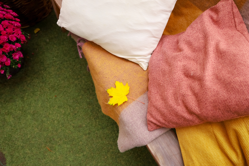 A yellow autumn leaf rests on pink, white, and orange cushions arranged on a bench with green grass and pink flowers nearby.