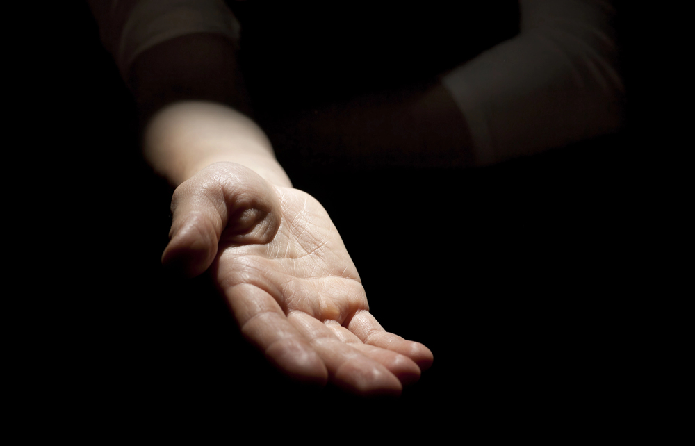 A single outstretched hand is illuminated against a dark background, palm facing upwards, suggesting a gesture of offering, asking, or openness.