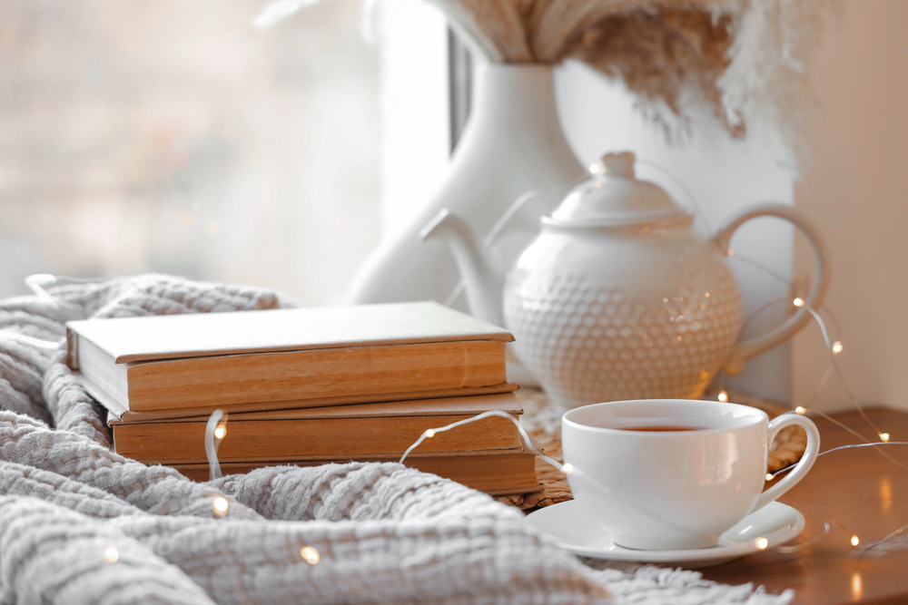 A cosy scene with two books, a white teacup and saucer, a white teapot, a soft knitted blanket, and warm fairy lights on a wooden surface near a window. Dried flowers are in a vase in the background.