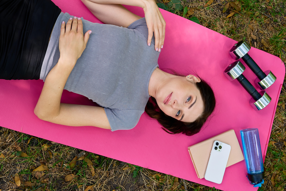 A woman lies on a pink exercise mat outdoors beside dumbbells, a water bottle, a mobile phone, and a notebook, surrounded by grass and fallen leaves.