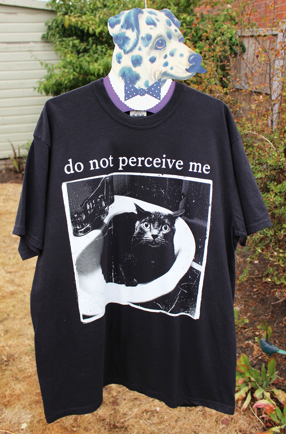 A black T-shirt with a photo of a cat in a bath and the text do not perceive me, displayed on a mannequin with a dog head mask in an outdoor garden setting.