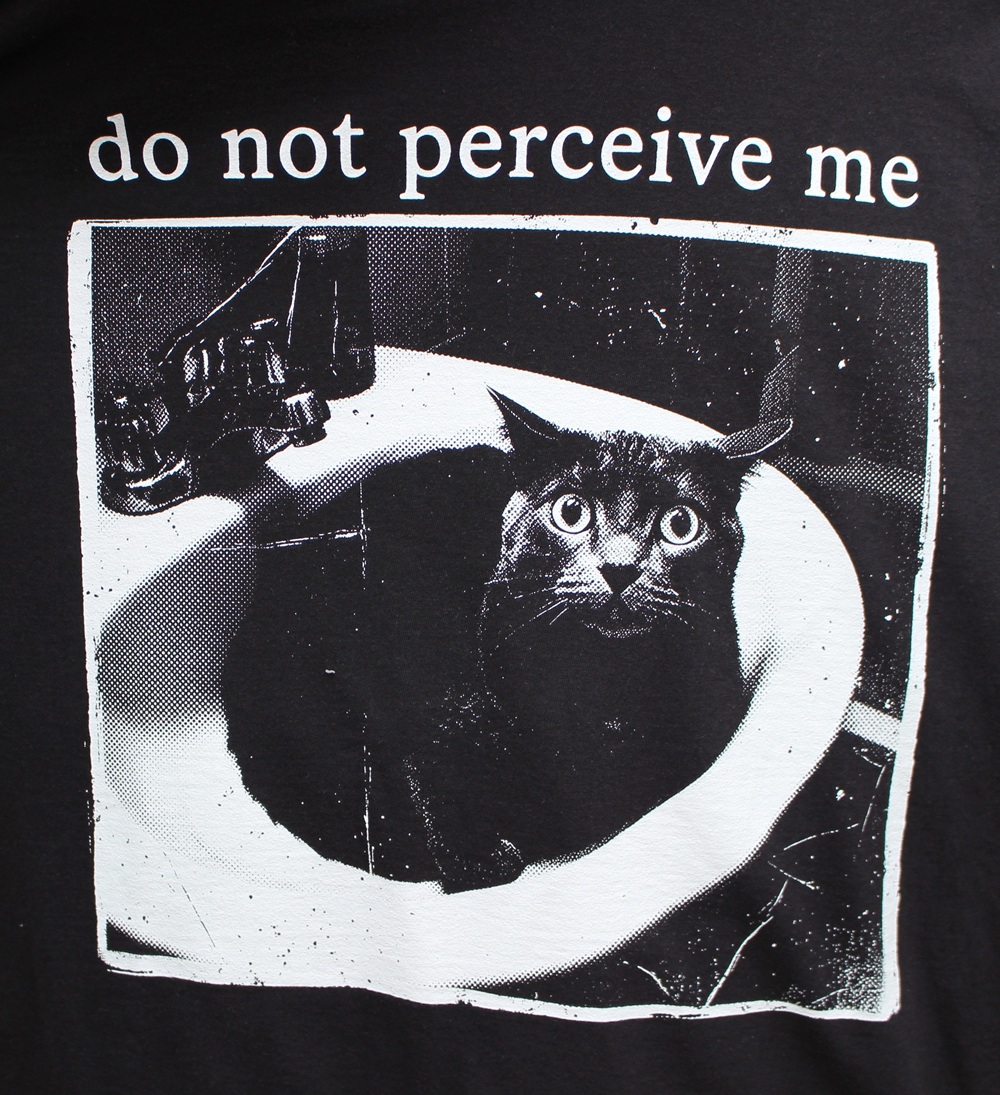 Black cat sitting in a white basin, looking wide-eyed. Text above reads, do not perceive me. The image appears on a black shirt with a high-contrast, graphic design style.