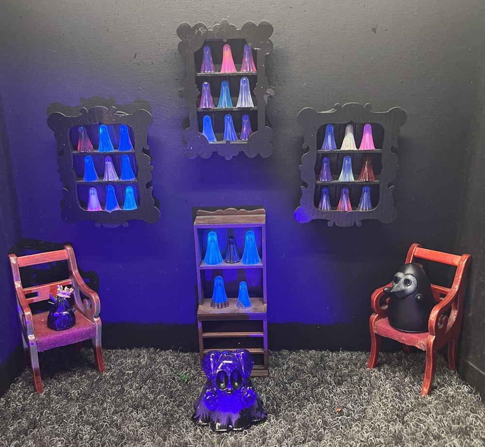 A small dark room with tiny shelves holding colourful ghost figurines, two red chairs with decorations, and a purple ghost figure on the floor. The scene has a playful, eerie vibe.