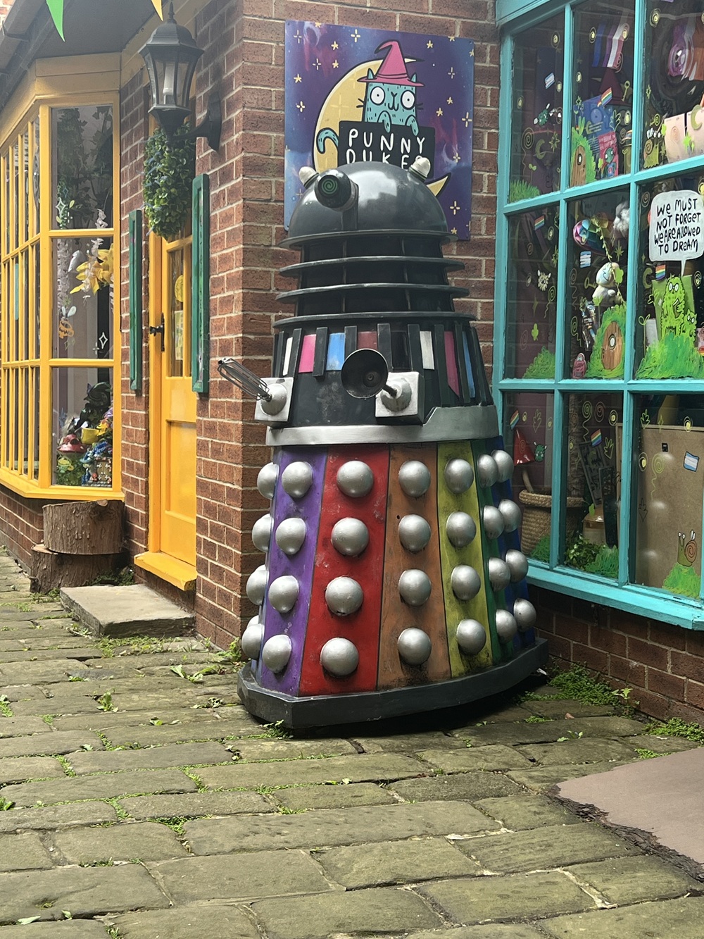 A colourful Dalek model stands outside a whimsical shop with bright yellow and blue doors, surrounded by playful decorations and artwork in the windows.