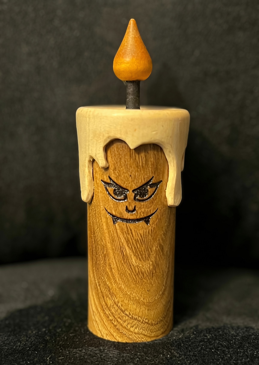 A wooden candle sculpture with a carved angry face, wax drips on top, and a small orange flame. The background is dark and out of focus.