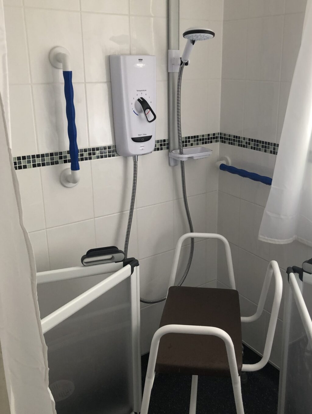 Accessible shower with a brown shower chair, grab rails, a handheld showerhead, a white shower control unit, and a privacy screen on the left side. White tiled walls with a black trim.