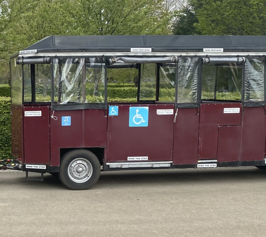 A maroon, accessible transport vehicle with wheelchair symbols on its side, clear plastic windows, and Mind the Step and Mind your Head warnings above and below the windows, parked on a paved path.
