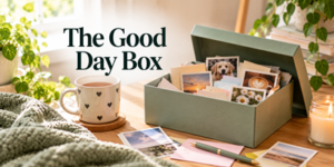 A green box filled with photos and cards sits on a wooden table next to a mug with hearts, a candle, a pen, and indoor plants. Text reads The Good Day Box. Sunlight streams into the cosy room.