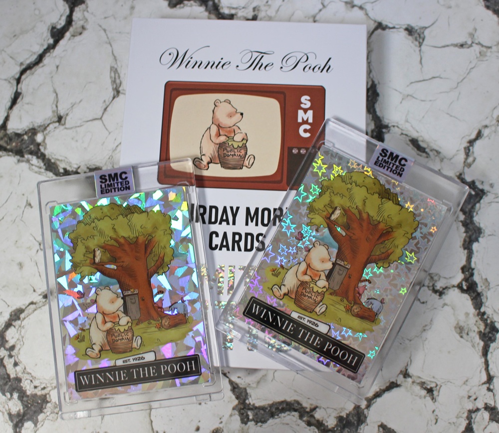 Two holographic Winnie the Pooh trading cards and a Saturday Morning Cards flyer are displayed on a white, textured surface. The cards show Pooh with honey under a tree, featuring reflective star patterns.