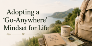 A travel scene with a notebook, compass, mug, and rucksack on a ledge overlooking nature. Text reads: Adopting a Go-Anywhere Mindset for Life..