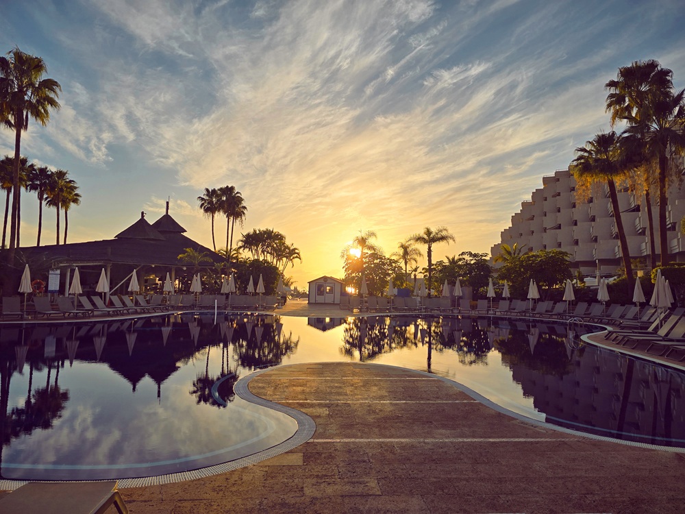 A swimming pool reflecting a sunset with palm trees, a resort building, and sun loungers in the background.