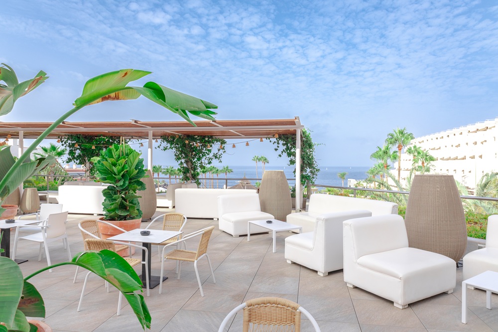 An outdoor terrace with white lounge seating, small tables, and potted plants overlooking the sea.