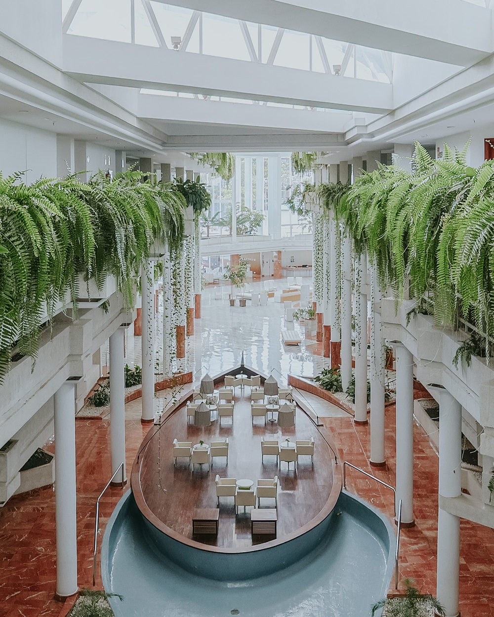 A boat-shaped platform with dining tables and chairs surrounded by a shallow pool in a large, airy atrium filled with hanging ferns.
