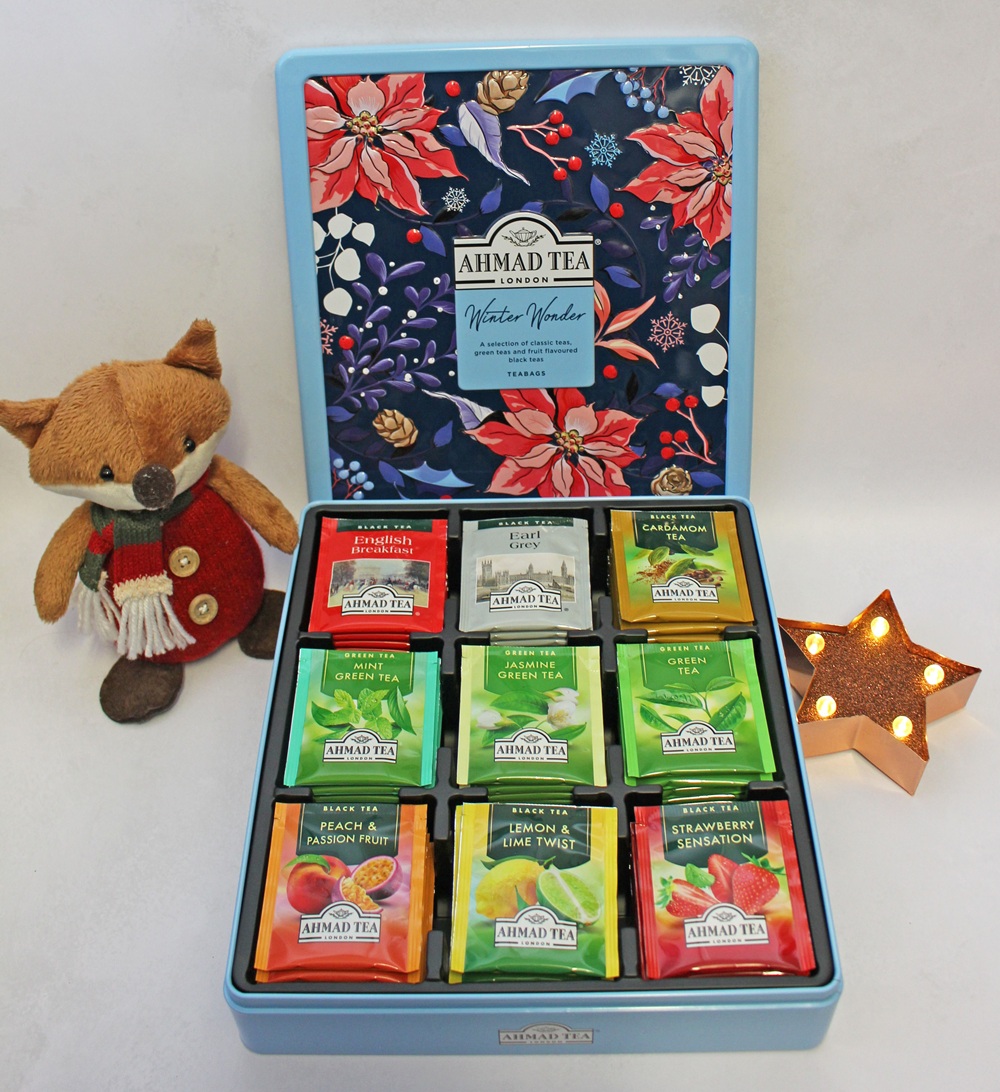 Open gift tin of Ahmad Tea Winter Wonder tea bags, featuring nine assorted flavours, next to a small plush fox and a lit star-shaped ornament.