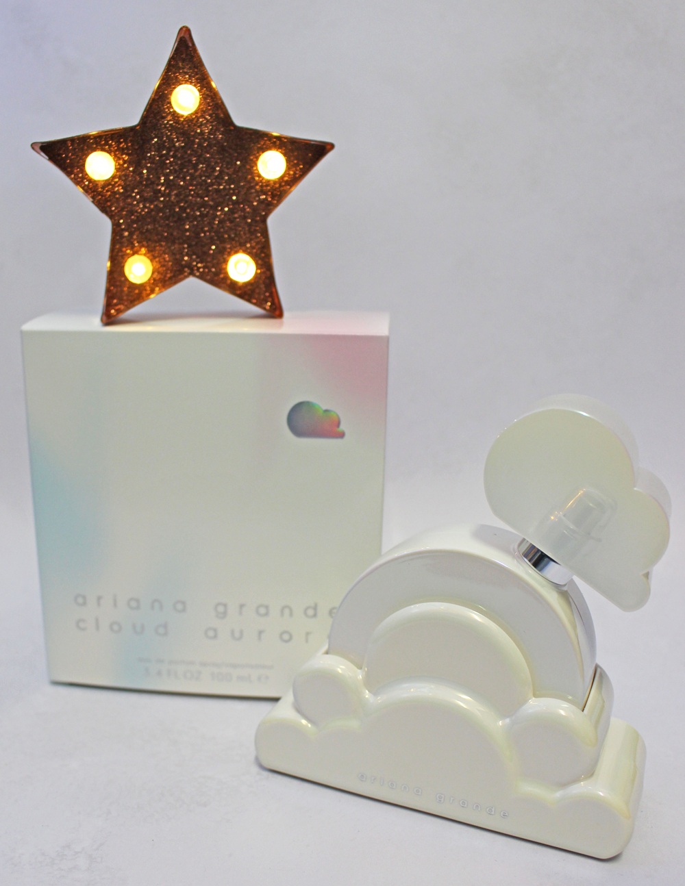Ariana Grande Cloud Aurora perfume bottle and box, with a lit, glittery star decoration behind them.