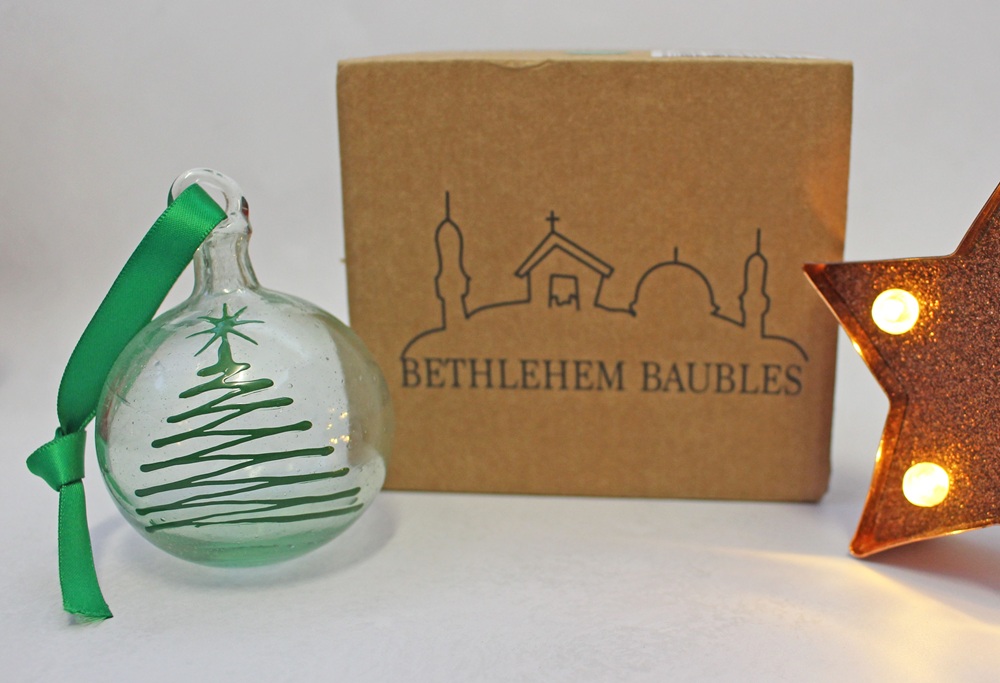 A clear glass Christmas bauble with a green ribbon and a green spiral tree design, positioned in front of a cardboard box labelled BETHLEHEM BAUBLES next to a lit star.