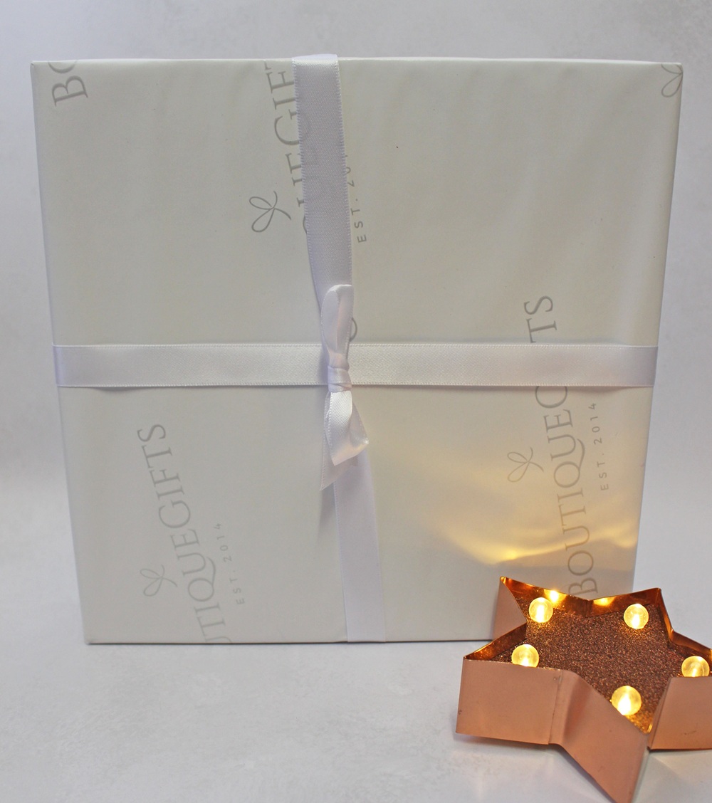 A square gift box wrapped in cream-coloured paper with the Boutique Gifts logo, tied with a white ribbon, sitting next to a small, illuminated star-shaped light.