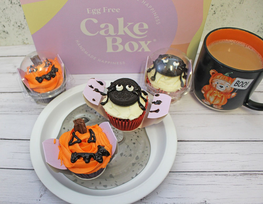 Three Hallowe’en-themed cupcakes with orange and white icing, positioned in front of an Egg Free Cake Box box and a mug of tea.