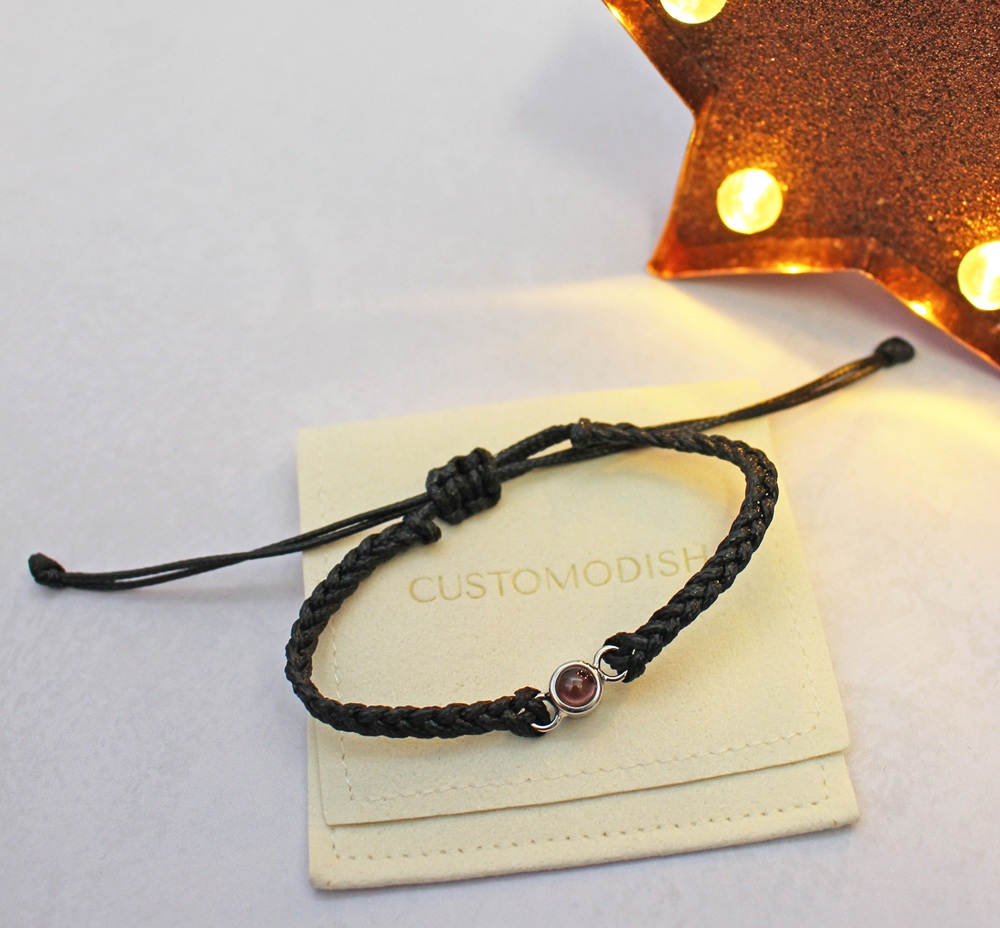 Black braided cord bracelet with a small, round, reddish-brown stone charm on a branded cream-coloured pouch.
