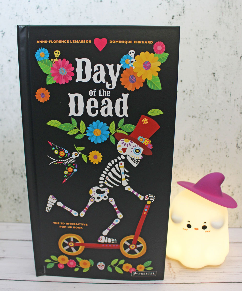 The cover of the pop-up book Day of the Dead featuring a skeleton on a scooter, displayed next to a small glowing ghost light wearing a purple witch’s hat.