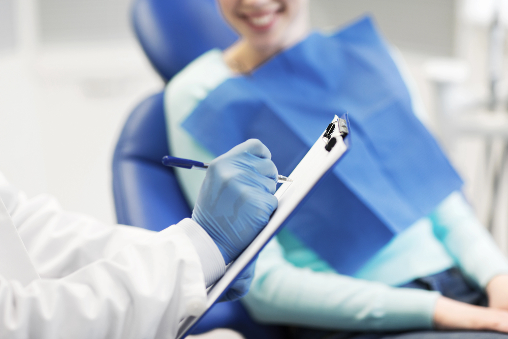 A dentist wearing blue gloves writes on a clipboard whilst a patient sits in a dental chair.