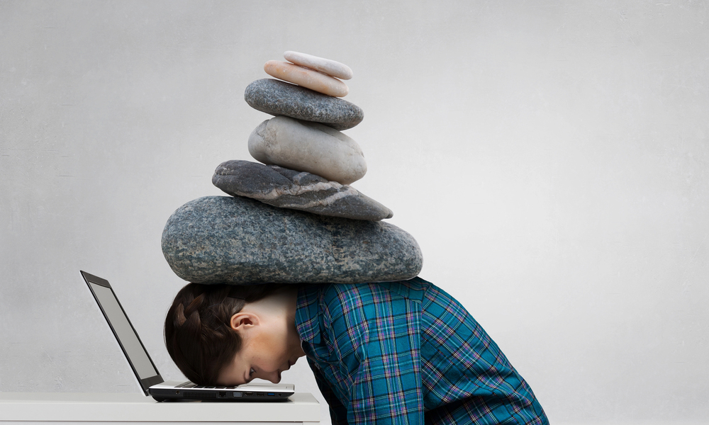 A person in a checked shirt rests their head on a laptop, with a tall, precarious stack of river stones balanced on their head.
