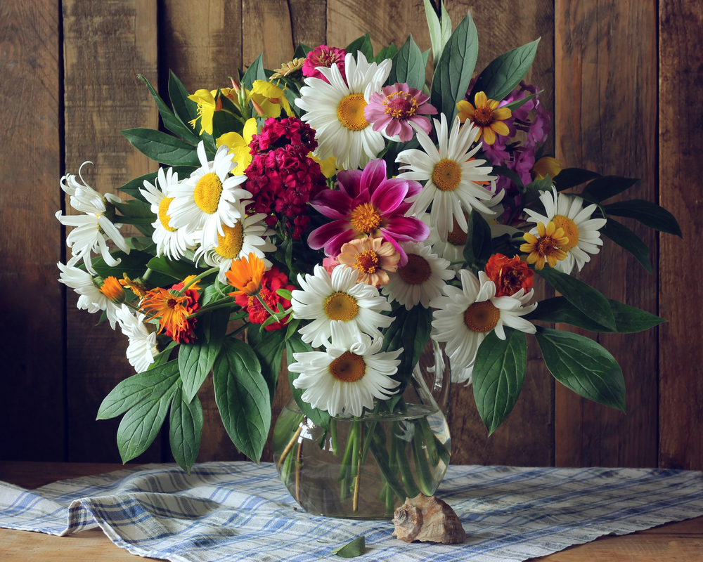 A vibrant floral bouquet featuring white daisies, pink dahlias, and other colourful flowers in a glass vase on a chequered cloth.