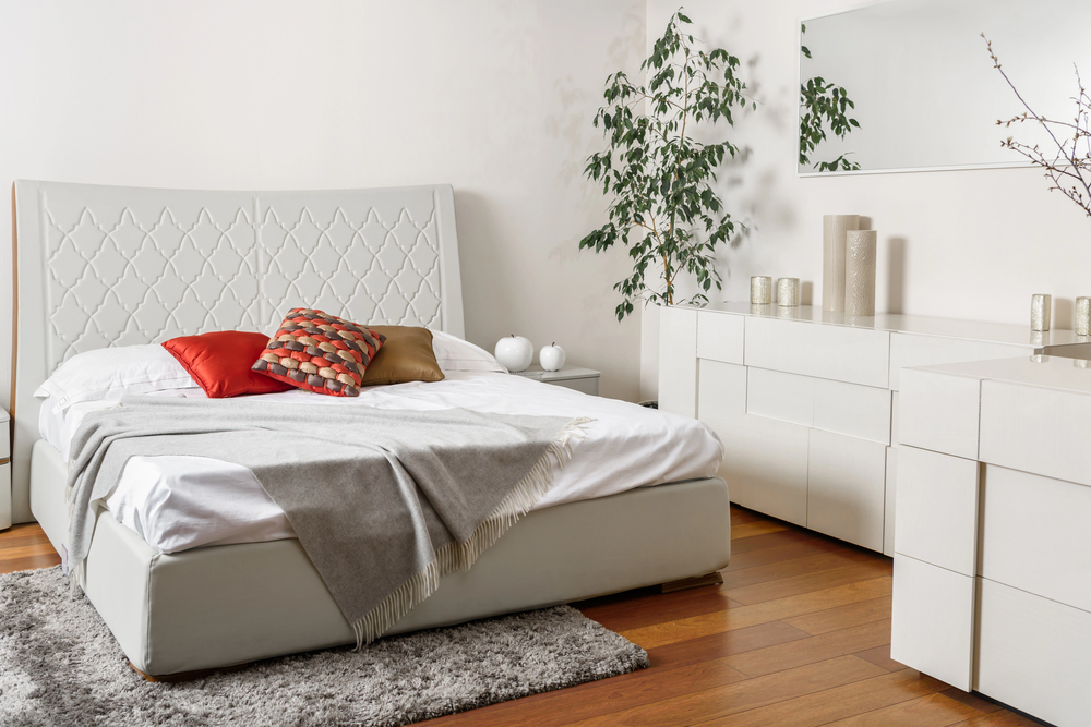 A bedroom with a white upholstered bed topped with red and brown cushions and a grey throw blanket, next to white storage cupboards and a tall plant.