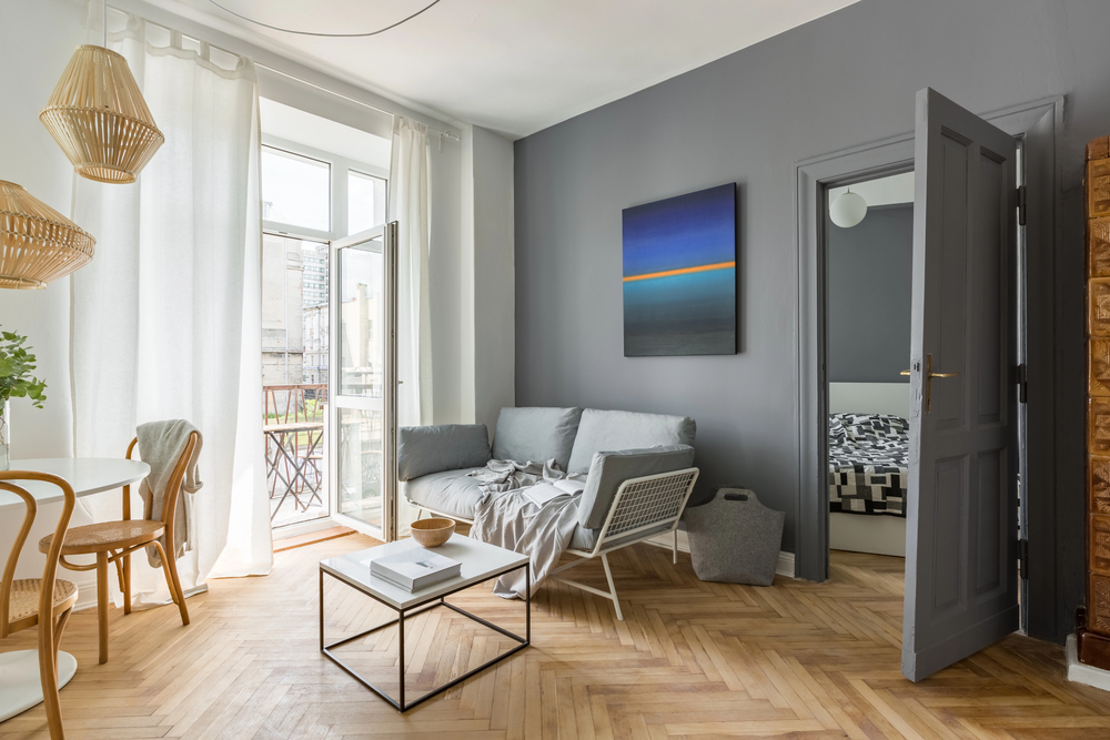 Living room with a grey sofa, a small white coffee table, and an abstract blue and orange painting on a grey wall, with an open doorway leading to a bedroom.