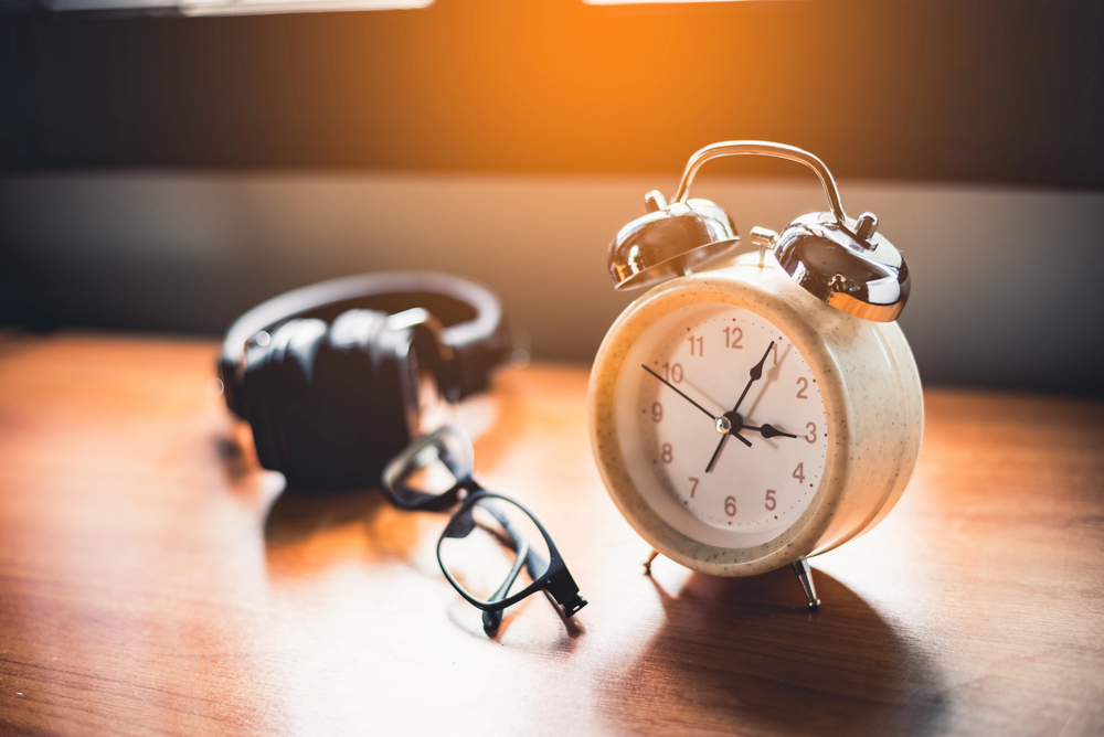 An analogue alarm clock, a pair of spectacles, and over-ear headphones on a wooden surface.
