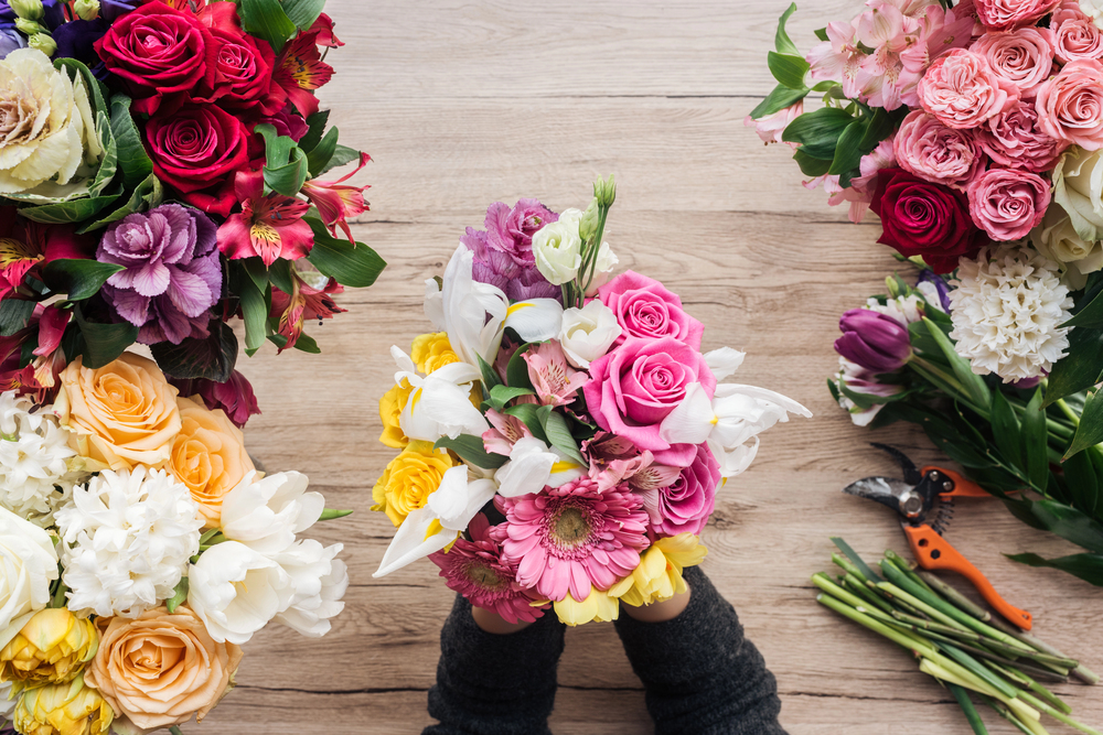 Hands hold a small, colourful bouquet of assorted flowers over a wooden table, surrounded by other flower arrangements and floral secateurs.