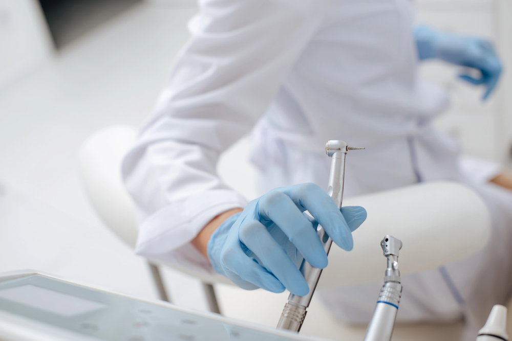A dentist wearing blue gloves holds a dental drill in their surgery.
