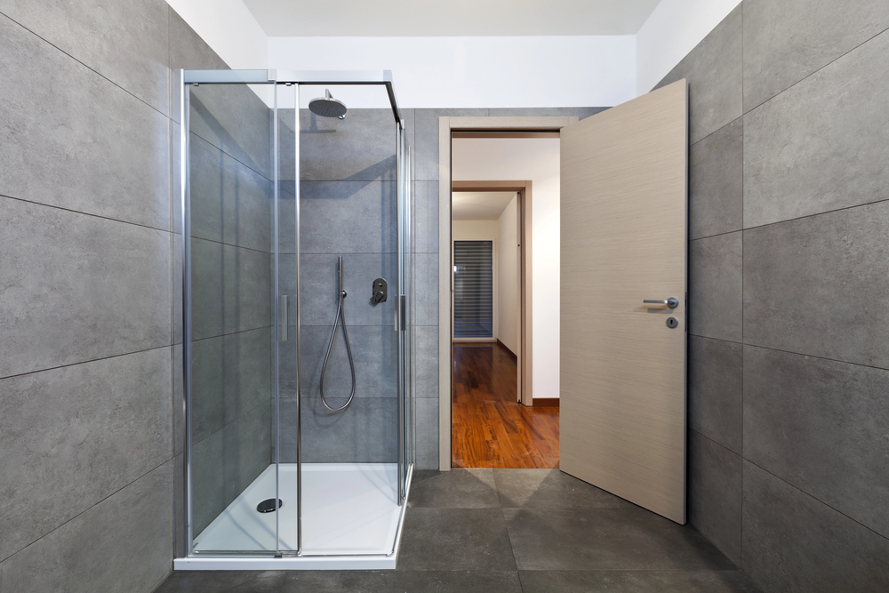 A modern shower with a glass enclosure stands in a bathroom with large grey tiled walls, next to an open doorway leading to a room with wooden flooring.