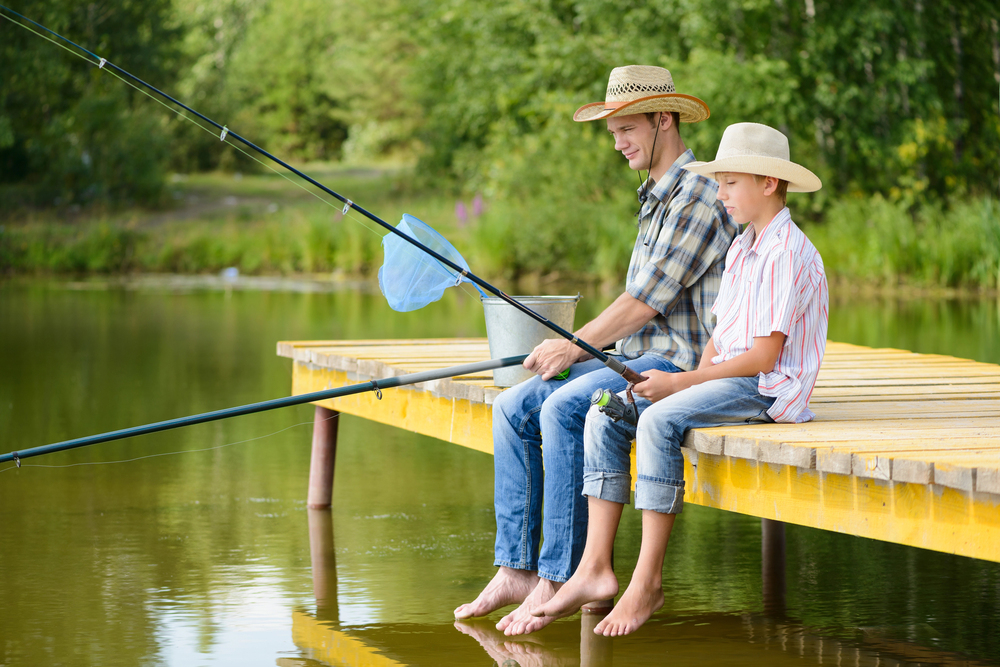 An adult and a boy wearing straw hats sit on a wooden jetty fishing in a lake.