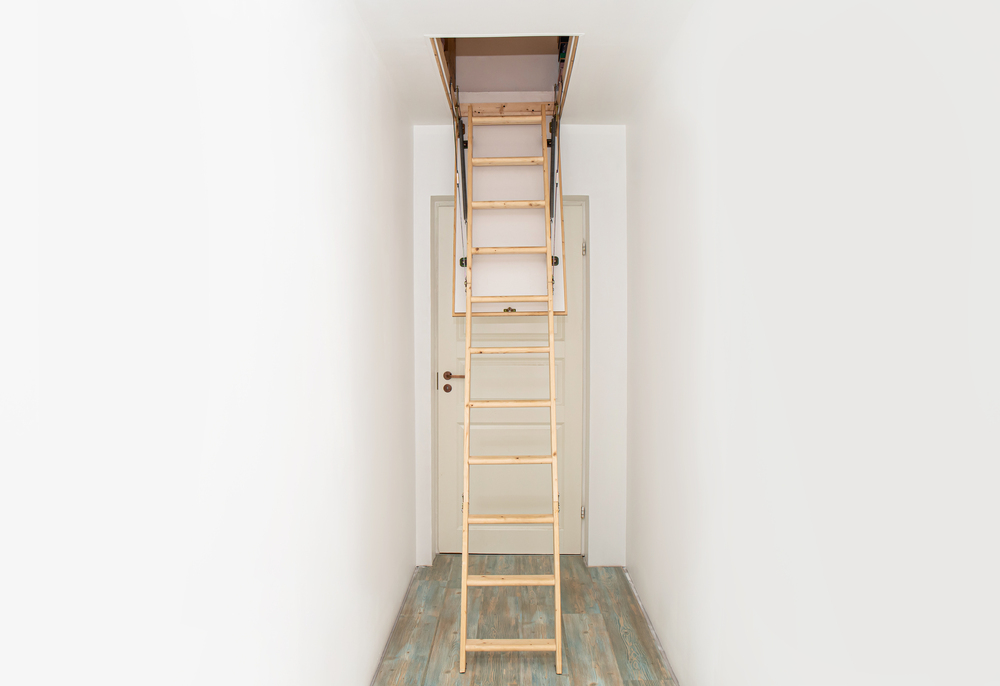 A wooden loft ladder unfolded from a ceiling hatch in a narrow corridor above a closed door.