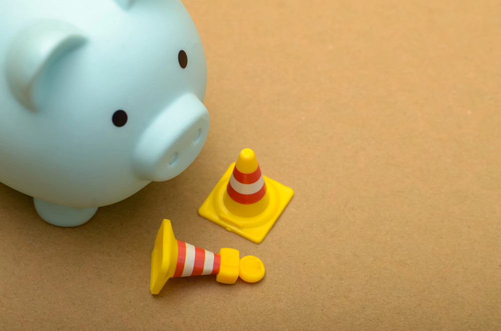 A light blue piggy bank sitting next to one upright and one fallen miniature orange traffic cone.