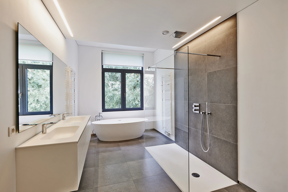 Modern bathroom with a double vanity unit, a freestanding oval bath in front of a window, and a walk-in shower with a glass partition and grey tiled walls.