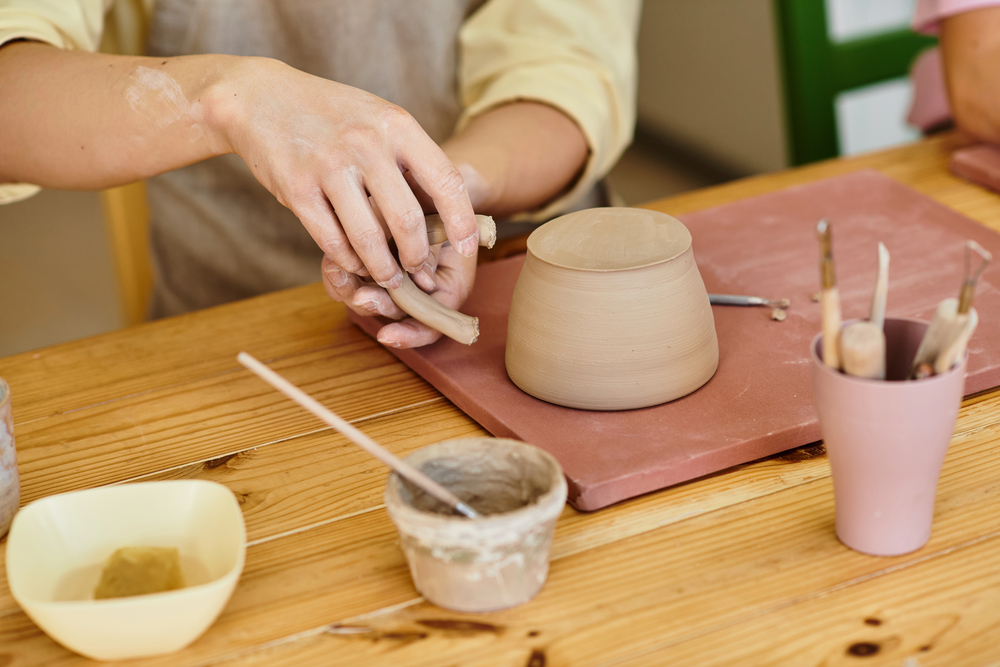 Hands attaching a clay handle to an inverted ceramic bowl on a wooden table with pottery tools.