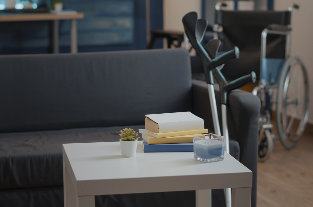 A small white table with books, a small succulent, and a candle, with crutches and a wheelchair in the background.