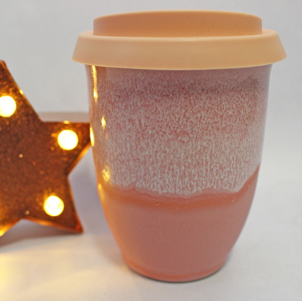 A pink ceramic travel mug with a light peach silicone lid, next to a lit star-shaped decoration.