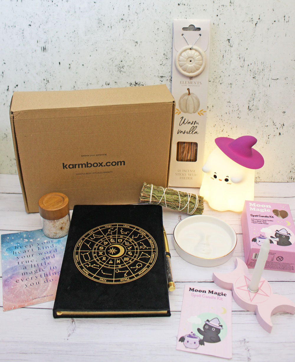 A Karmbox subscription box surrounded by a zodiac-themed diary, a ghost-shaped lamp, incense, a spell candle kit, and small ritual items.