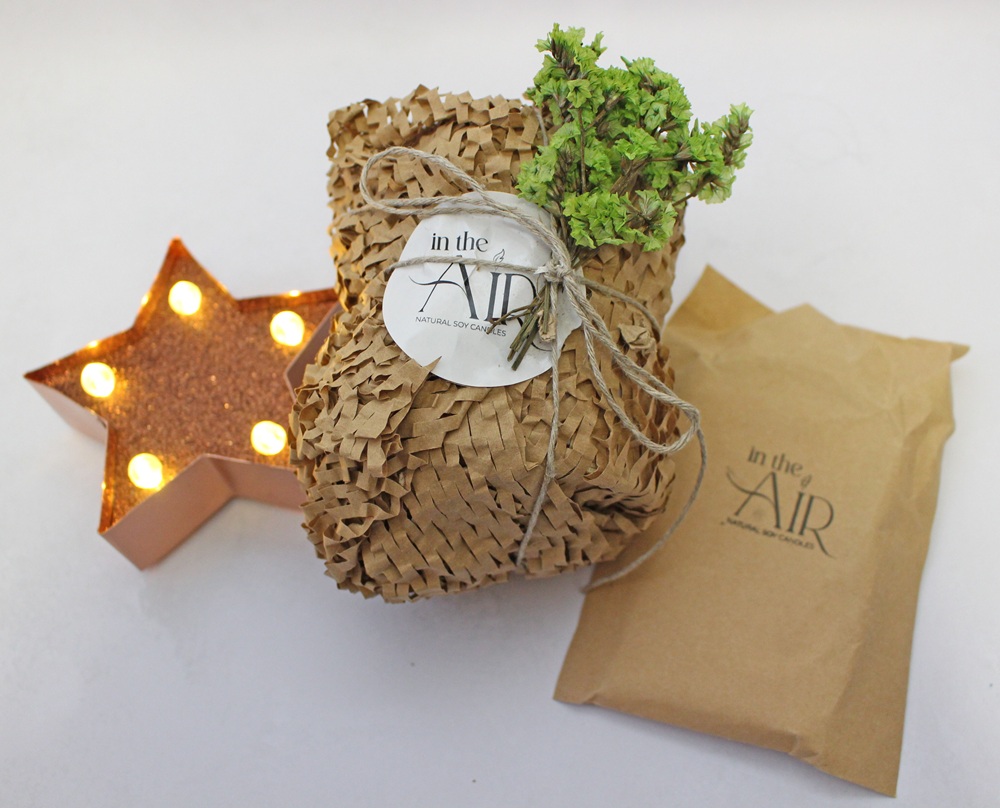 A candle wrapped in brown honeycomb paper with an In the Air branded tag and a sprig of greenery, next to a matching branded pouch and a lit copper-coloured star.