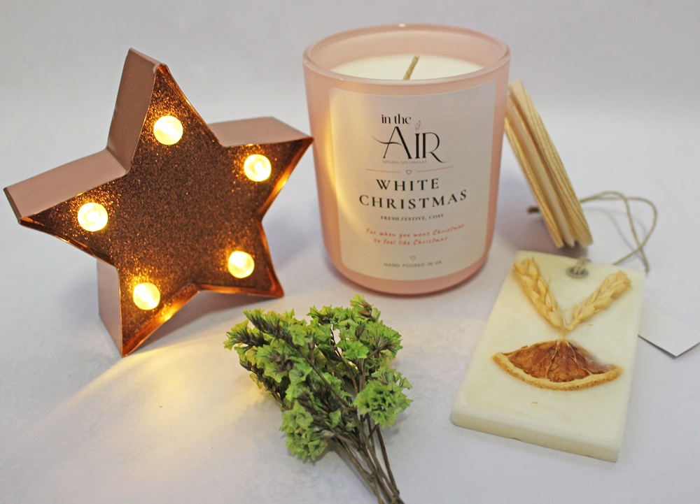 A lit star-shaped lamp next to a White Christmas soy candle, a wax melt decorated with dried orange and wheat, and a small bunch of green herbs.